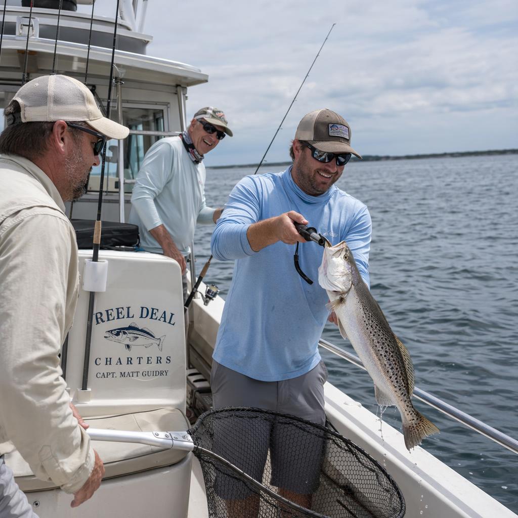 Charter fishing at Holdne Beach - Book direct. Skip the fees. Man on a fishing boat holds up a striped bass with pliers, smiling as two friends watch near the rail. Sign reads 'Reel Deal Charters' on the boat side.
