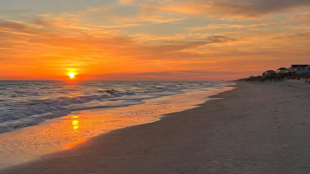 Holden Sunset - Book direct. Skip the fees. Sunset over a sandy beach with orange skies, reflecting on wet sand as gentle waves roll in, with houses along the right shoreline.